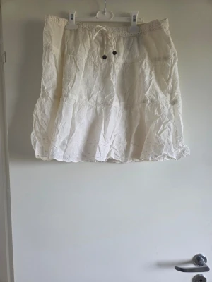 Vit kortkjol med broderade detaljer, stl 42 - Söt vit kortkjol med broderade detaljer och snörning i midjan. Kjolen har en luftig och lätt känsla, perfekt för varma dagar. Volangkant nedtill ger en extra feminin touch. Passar till många olika stilar och är enkel att matcha.