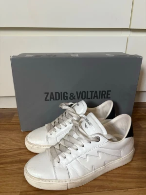 Vita sneakers från Zadig & Voltaire - Snygga vita sneakers från Zadig & Voltaire med cool blixtdetalj på sidan och svart hälparti. Skorna har vita snören, silverfärgade öljetter och är tillverkade i läder. Perfekta för dig som gillar stilrena men ändå unika sneakers. Storlek 37 men något större skulle jag säga. Nypris köpta på Zalando för 3095:- De är i väldigt bra skick och tvättas självklart innan fraktning. 
