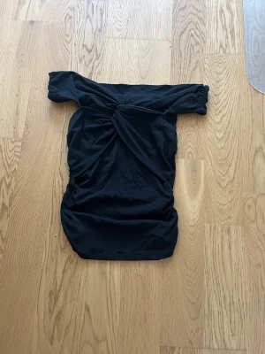 Svart offshoulder topp - Trendig svart topp med offshoulder och twistad detalj framtill. Toppen har en tight passform och är tillverkad i ett mjukt, stretchigt material som formar sig efter kroppen. Perfekt för dig som gillar en stilren och modern look.