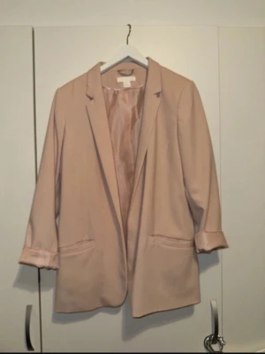 Ljusrosa oversized kavaj - Snygg ljusrosa kavaj med avslappnad, oversized passform och klassisk krage. Kavajen har två fickor framtill och uppvikta ärmslut med blankt foder som syns. Materialet ser ut att vara en mjuk polyesterblandning. Perfekt för dig som gillar stilrena och trendiga plagg.