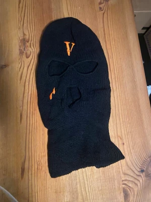 V-Lone Svart stickad balaclava  - Säljer en svart stickad balaclava med broderade orange detaljer, bland annat ett V på pannan. Masken har hål för ögon och mun. Perfekt för dig som vill ha en unik och edgy accessoar till vintern.
