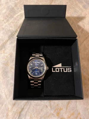 Lotus klocka  - Stilren klocka från Lotus med silverfärgad länkarmband och blå urtavla. Ny pris 1300kr. Den är i ett jättebra skick, knappt använd. Klockan har romerska siffror, datum- och veckovisning samt markerad boett. Perfekt accessoar för dig som gillar klassisk men modern stil. 