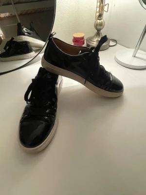 Morris lady  - Snygga svarta sneakers i glansigt lackmaterial med svarta skosnören och vita, tjocka gummisulor. Låg modell som passar perfekt till streetwear och casual outfits. Insidan är beige och de har en klassisk rund tå.