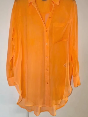 Orange skjorta från DKNY - Snygg orange skjorta från DKNY i transparent och lätt material. Skjortan har lång ärm, klassisk krage, bröstficka och knappar framtill. Perfekt för dig som gillar färg och vill sticka ut lite extra. Tvättlappen är numera bortklippt. 