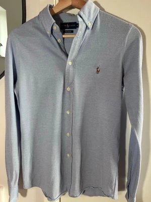 Blå skjorta från Ralph Lauren - Säljer en jättefin ljusblå långärmad oxfordskjorta från Polo Ralph Lauren i modellen "Knit Oxford." Materialet är mjukt och har lite stretch, vilket gör den superbekväm att bära jämfört med en vanlig skjorta.  • Färg: Ljusblå. • Material: 100% Bomull (Knit Oxford/Piké-struktur). • Märke: Polo Ralph Lauren. • Storlek: Medium men sitter som Small • Nypris: Ca 1 400 kr. • Skick: Mycket bra skick