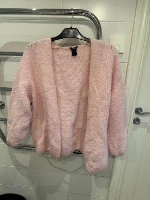 Ljusrosa kofta från Lindex - Mysig ljusrosa stickad kofta från Lindex med öppen front och avslappnad passform. Koftan har långa ärmar och är tillverkad i ett mjukt, fluffigt material som känns skönt mot huden. Perfekt att slänga över en t-shirt eller topp för extra värme. Storlek M/L