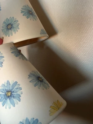 Blommiga lampskärmar i tyg - Två lampskärmar i vitt tyg med tryckta blå och gula blommor. Skärmarna har en klassisk konformad design och ger ett retroinspirerat intryck. Perfekt för att ge rummet en färgglad och personlig touch.