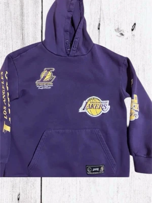 Lila Lakers hoodie med tryck - Snygg lila hoodie med Los Angeles Lakers tryck och broderade loggor på bröstet och ärmen. Stor ficka framtill och huva. Gula detaljer och text längs ena ärmen. Perfekt för dig som älskar basket och streetwear.