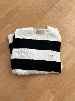Randig stickad tröja från GANT - Säljer en snygg kabelstickad tröja från GANT med breda marinblå och vita ränder. Tröjan har rund halsringning, ribbade muddar och en liten broderad logga på bröstet. Perfekt för dig som gillar klassisk preppy stil.