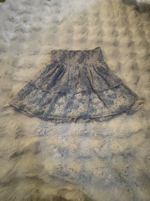 Blommig volangkjol med smock - Supersöt kjol med blått blommönster på vit botten och bred smock i midjan. Kjolen har fina volanger. Perfekt för dig som gillar en feminin och somrig stil.