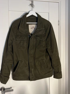 Olivgrön jacka från Hollister - Snygg olivgrön jacka från Hollister som är i storlek L men sitter lite mer mot M hållet. med fyra fickor framtill, knappar och hög krage. Jackan har en klassisk, rak passform och är tillverkad i bomullsmaterial. Perfekt för dig som vill ha en stilren och enkel look.