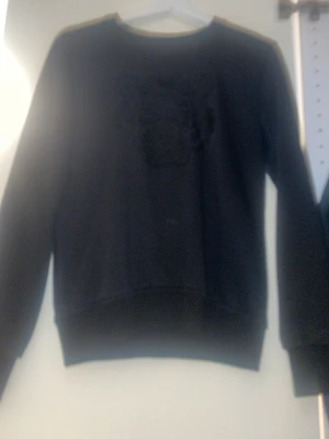 Svart sweatshirt från Kenzo - Säljer en svart sweatshirt från Kenzo med det ikoniska tigerhuvudet broderat på bröstet. Tröjan har rund halsringning, långa ärmar och ribbade muddar. Tillverkad i mjukt bomullsmaterial som är skönt att bära. Perfekt för dig som gillar streetwear och stilrena detaljer.