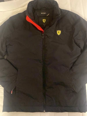 Svart pufferjacka från Ferrari - Snygg svart pufferjacka från Ferrari med gul Ferrari-logga på bröstet och cool röd detalj på insidan av kragen. Jackan har dragkedja framtill och två sidofickor. Perfekt för dig som vill ha en sportig och varm vinterjacka med exklusiv känsla.