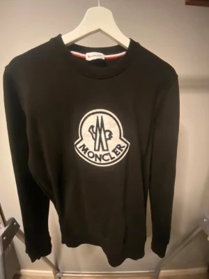 Svart sweatshirt från Moncler - Svart sweatshirt från Moncler med stort vitt broderat Moncler-logo på bröstet. Klassisk rund halsringning och långa ärmar. Perfekt för dig som gillar streetwear och vill ha en stilren look.