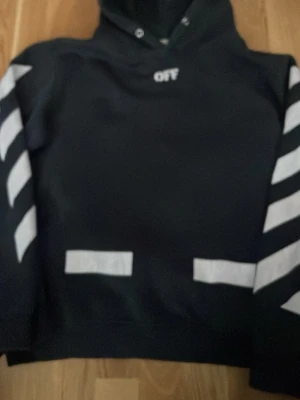 Svart Off-White hoodie med vita detaljer - Säljer en svart hoodie från Off-White med vita diagonala ränder på ärmarna och vita tryck på framsidan. Klassisk huva med dragsko och Off-White-logga tryckt vid halsen. Perfekt för dig som gillar streetwear och vill sticka ut. Den är lite solbränd 