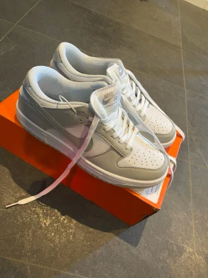 Nike Dunk Low vit/grå sneaker - Nike Dunk Low sneakers i vitt och ljusgrått läder. Klassisk låg siluett med perforerad tåbox, vita skosnören och ikoniska Swoosh-loggan på sidan. Sitter på en vit sula med grå detaljer. Perfekt för dig som gillar stilrena och tidlösa sneakers.