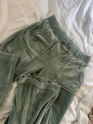 Gröna velourbyxor Juicy Couture - Mysiga gröna mjukisbyxor från Juicy Couture i mjuk velour. Byxorna har bred resår i midjan med dragsko, stora fickor fram och bak med knappdetaljer samt broderad logga på ena fickan. Perfekta för chill dagar och en avslappnad stil.