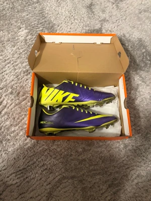 Nike Mercurial lila/gula fotbollsskor - Säljer ett par Nike Mercurial fotbollsskor i lila och gult med stora Nike-loggor på sidan. Skorna har snörning, låg profil och dobbar för spel på gräs. Tillverkade i syntetmaterial för lätt vikt och bra bollkontroll. Perfekta för dig som vill sticka ut på planen.