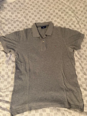 Grå pikétröja från Gant - Klassisk grå pikétröja från Gant med liten broderad logga på bröstet. Tröjan har korta ärmar, krage och knappar framtill. Perfekt till jeans eller shorts för en clean och enkel stil. OBS! Finns en liten knappa vit fläck under armhålorna