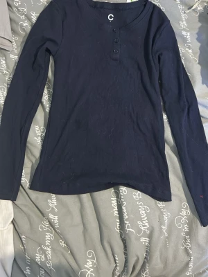 Svart ribbad långärmad topp - En svart långärmad topp med ribbad struktur och rund halsringning. Toppen har knappar framtill och är i en mjuk, stretchig bomullsblandning. Enkel och stilren modell som passar till mycket.