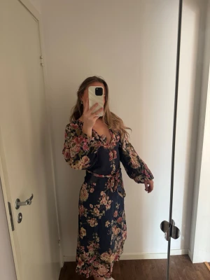 Marinblå blommig omlottklänning - Säljer en marinblå långklänning med blommigt mönster. Klänningen har omlottmodell med v-ringning, långa ärmar och ett matchande knytband i midjan. Perfekt för dig som gillar romantisk och bohemisk stil. Nypris 999kr