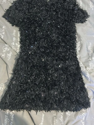 Svart paljettklänning med kort ärm - Säljer en svart kortklänning med korta ärmar, täckt av glittriga paljetter och fluffig textur. Klänningen har en rak passform och är perfekt för dig som vill sticka ut. Materialet känns syntetiskt och ger en cool look.