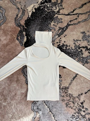 Vit/beige ribbad polotopp med cut out - Trendig vit/beige ribbad polotopp med lång ärm och cool cut out-detalj vid axeln. Polokrage ger en snygg siluett och materialet är stretchigt och mjukt. Perfekt för dig som vill sticka ut lite extra. Anledningen till att jag säljer är på grund av att jag har tröttnat på den, däremot så är den otroligt vacker. 