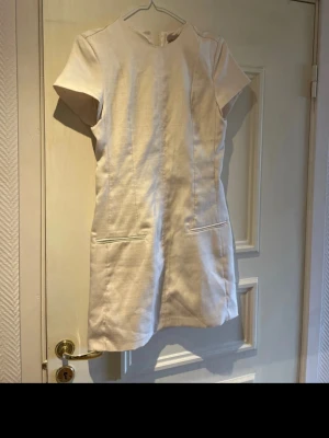 Ljus kort klänning från H&M - Kort, ljus klänning från H&M med enkel och stilren design. Klänningen har korta ärmar, rund halsringning och två diskreta fickor framtill. Materialet har en lätt struktur och klänningen stängs med dragkedja i ryggen.