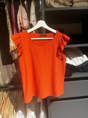 Orange topp med volangärmar från Longboard - Säljer en snygg orange topp från Longboard med volangdetaljer på ärmarna. Toppen har rund halsringning och är tillverkad i ett strukturerat tyg som ger en cool känsla. Perfekt för dig som vill sticka ut med färg och stil.