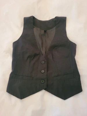 Mörkgrå kostymväst med knappar - Snygg mörkgrå kostymväst med tre knappar framtill och två dekorativa fickor. Västen har en klassisk V-ringning och ett randigt innerfoder. Perfekt att styla över en skjorta för en dressad look.