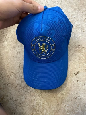 Blå Chelsea FC keps - Snygg blå keps med Chelsea FC:s officiella klubbmärke framtill i guld och blått. Justerbar rem baktill för perfekt passform. Tillverkad i ett slitstarkt syntetmaterial som andas. Perfekt för dig som vill visa ditt stöd för Chelsea på ett stilrent sätt.