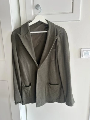 Olivgrön kavaj med fickor - Stilren olivgrön kavaj med två framfickor och klassisk krage. Kavajen har enkelknäppning och är tillverkad i ett mjukt, strukturerat tyg. Perfekt för dig som vill ha en avslappnad men ändå dressad look.