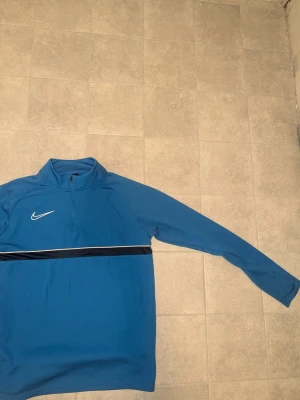 Blå Nike Dri-FIT träningströja - Snygg blå långärmad träningströja från Nike med Dri-FIT-material som andas. Tröjan har halv dragkedja framtill, vit Nike-logga på bröstet och en svart/vit rand över bröstet. Perfekt för träning eller uppvärmning.