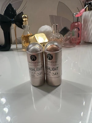 Ronassi Highlight Stick 3D Duo - Två highlighter sticks från Ronassi i 3D-utförande. Ena sticken har en kall, silvrig ton och den andra en varm, champagneguldig nyans. Båda kommer i smidiga stift med genomskinliga lock, perfekta för att skapa glow på kindben och brynben.