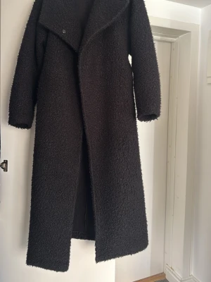 Lång svart teddyjacka med bälte - Säljer en lång svart teddyjacka med bred krage. Jackan har ett matchande bälte i midjan och är gjord i mjukt, fluffigt teddy-material. Perfekt för att hålla sig varm och snygg under kalla dagar.