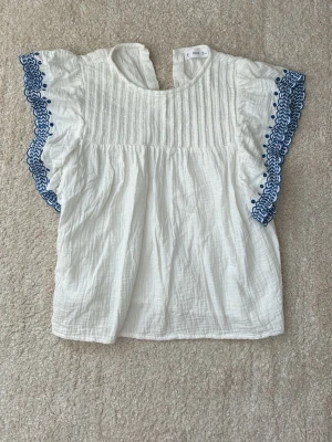 Vit blus med blå brodyr  - Supersöt vit blus med volangärmar och blå broderade detaljer längs kanterna. Blusen har rund halsringning och är gjord i ett lätt, luftigt bomullstyg med struktur. Perfekt för en fräsch och somrig look!