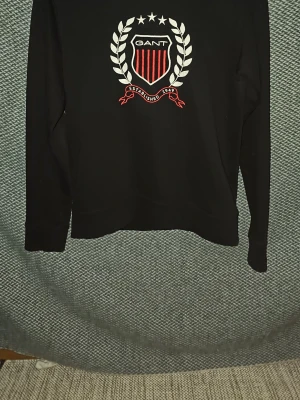 Svart sweatshirt från Gant - Svart sweatshirt från Gant med broderad logga framtill. Loggan har vita och röda detaljer, texten 'Established 1949' och ett sköldmärke med stjärnor och lagerkrans. Tröjan har rund hals och långa ärmar. Perfekt för en sportig och stilren look. Är öppen för prisförslag vid snabb försäljning.
