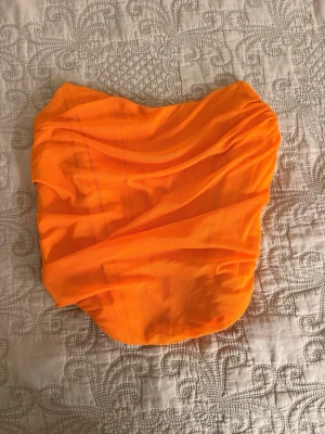 Orange bandeau tubtopp med dragkedja - Snygg orange bandeau tubtopp med draperad mesh och dragkedja bak. Toppen är axelbandslös och har en cool, croppad passform som passar perfekt till höga jeans eller kjol. Materialet känns lätt och luftigt, perfekt för varma dagar.