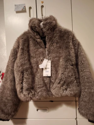 Grå fluffig fuskpälsjacka Zara - Supersnygg grå fuskpälsjacka med hög krage och mjuk, fluffig känsla. Jackan har en rak passform och stängs framtill. Perfekt för dig som vill ha en trendig och varm look under vintern. Märke och storlek framgår ej.