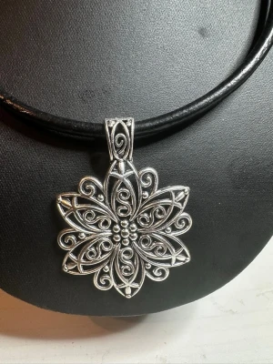 Svart halsband med silverfärgat blom-hänge - Unikt halsband med svart läderrem och stort, detaljerat blomformat hänge i silverfärgad metall. Hänget har fina snirkliga mönster och små pärlliknande detaljer i mitten. Perfekt för dig som vill sticka ut med en cool accessoar.
