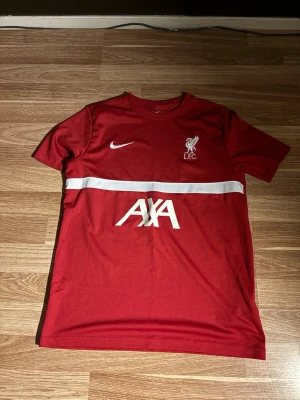 Liverpool FC röd träningströja Nike - Säljer en röd Liverpool FC träningströja från Nike med vit logga och sponsortryck. Tröjan har rund hals, korta ärmar och en vit horisontell rand över bröstet. Perfekt för fotbollsträning eller att bära till vardags.