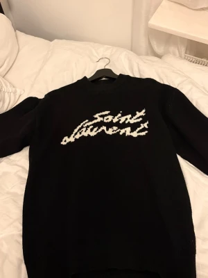Svart stickad Saint Laurent tröja - Cool svart stickad tröja från Saint Laurent med vit logotyp i ett handskrivet typsnitt på bröstet. Tröjan har rund hals och långa ärmar, perfekt för att ge en trendig look. Materialet ser ut att vara mjukt och värmande.