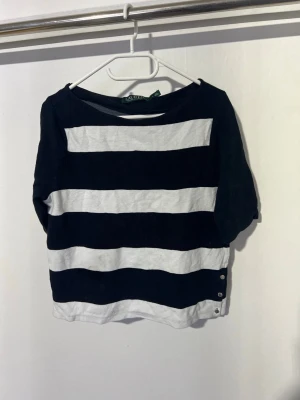 Randig topp från Ralph Lauren - Säljer en svartvit randig topp från Ralph Lauren med breda horisontella ränder. Toppen har trekvartsärm och knappar på sidan nedtill för extra detalj. Perfekt för en avslappnad och stilren look.