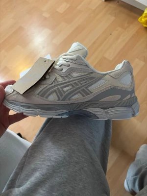 Vita och grå ASICS sneakers - Snygga sneakers från ASICS i vitt och ljusgrått med klassiska detaljer och logga på sidan. Skorna har en chunky sula och är tillverkade i mesh och syntetmaterial för en sportig vibe. Perfekta för dig som gillar stilrena och bekväma sneakers.