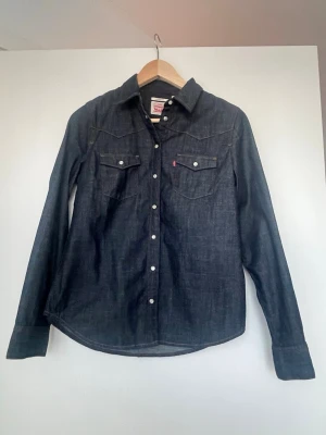 Mörkblå jeansskjorta från Levi's - Säljer en klassisk mörkblå jeansskjorta från Levi's med tryckknappar och två bröstfickor. Skjortan har lång ärm och en snygg western-inspirerad design. Perfekt för dig som gillar tidlös denimstil.