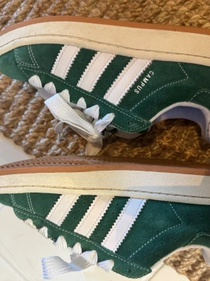 Adidas Campus gröna sneakers - Gröna Adidas Campus sneakers  med klassiska vita ränder på sidorna och vit sula. Skorna har breda vita skosnören och gummisula i brunt. Ikonisk retrostil med låg profil