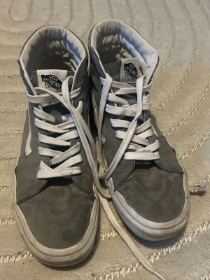 Ljusblåa/gråa Vans High sneakers - Ljusblåa/gråa Vans High sneakers med vit sula och vita skosnören. Klassisk hög modell med ikonisk sidostripes i vitt och vadderad krage för extra komfort. Ovandelen är i mocka och textil, perfekt för en streetwear-look.