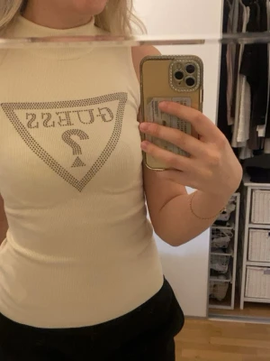 Beige ribbad linnetopp från Guess - Säljer en beige ribbad linnetopp från Guess med hög hals och ikonisk logga framtill i silverfärgade nitar. Toppen är ärmlös och har en tight passform. Är bara använd några gånger så skicket är fint.