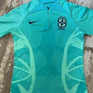  Brasilien träningströja - Snygg turkos träningströja från Nike med Brasilien-logga och modern grafisk design. Tröjan har korta ärmar, halv dragkedja och meshdetaljer för extra ventilation. Perfekt för fotbollsträning eller sportiga aktiviteter. Priset är ej hugget i sten!