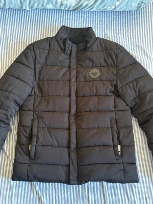 Svart pufferjacka från Armani Exchange - Snygg svart pufferjacka från Armani Exchange med diskret logomärke på bröstet. Jackan har ståkrage, två sidofickor med dragkedja och quiltad design. Perfekt för kalla dagar och ger en clean look. Litet hål bakom jackan.
